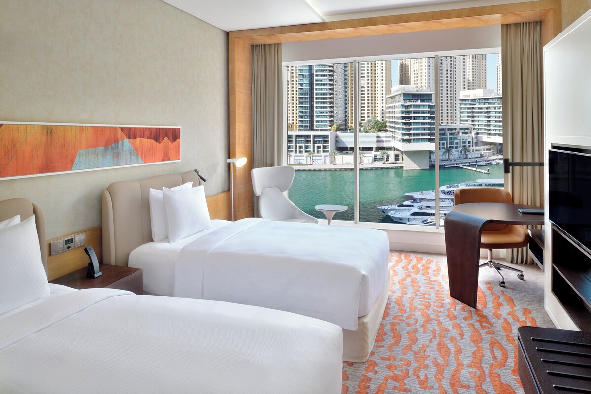 Une chambre d’hôtel avec deux lits, un bureau, une télévision et une grande fenêtre donnant sur un port de plaisance et des immeubles.