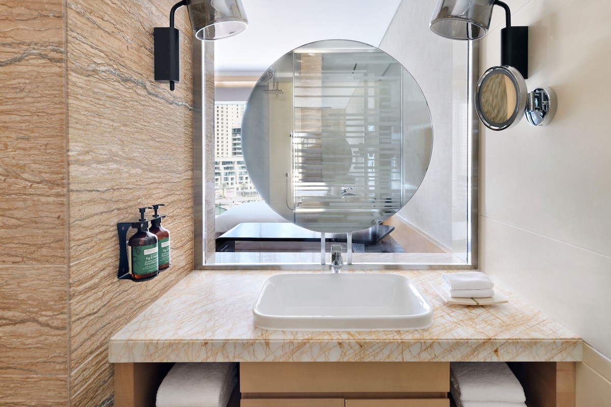 A modern bathroom with a marble countertop, a round mirror, a sink, and two wall-mounted lights.