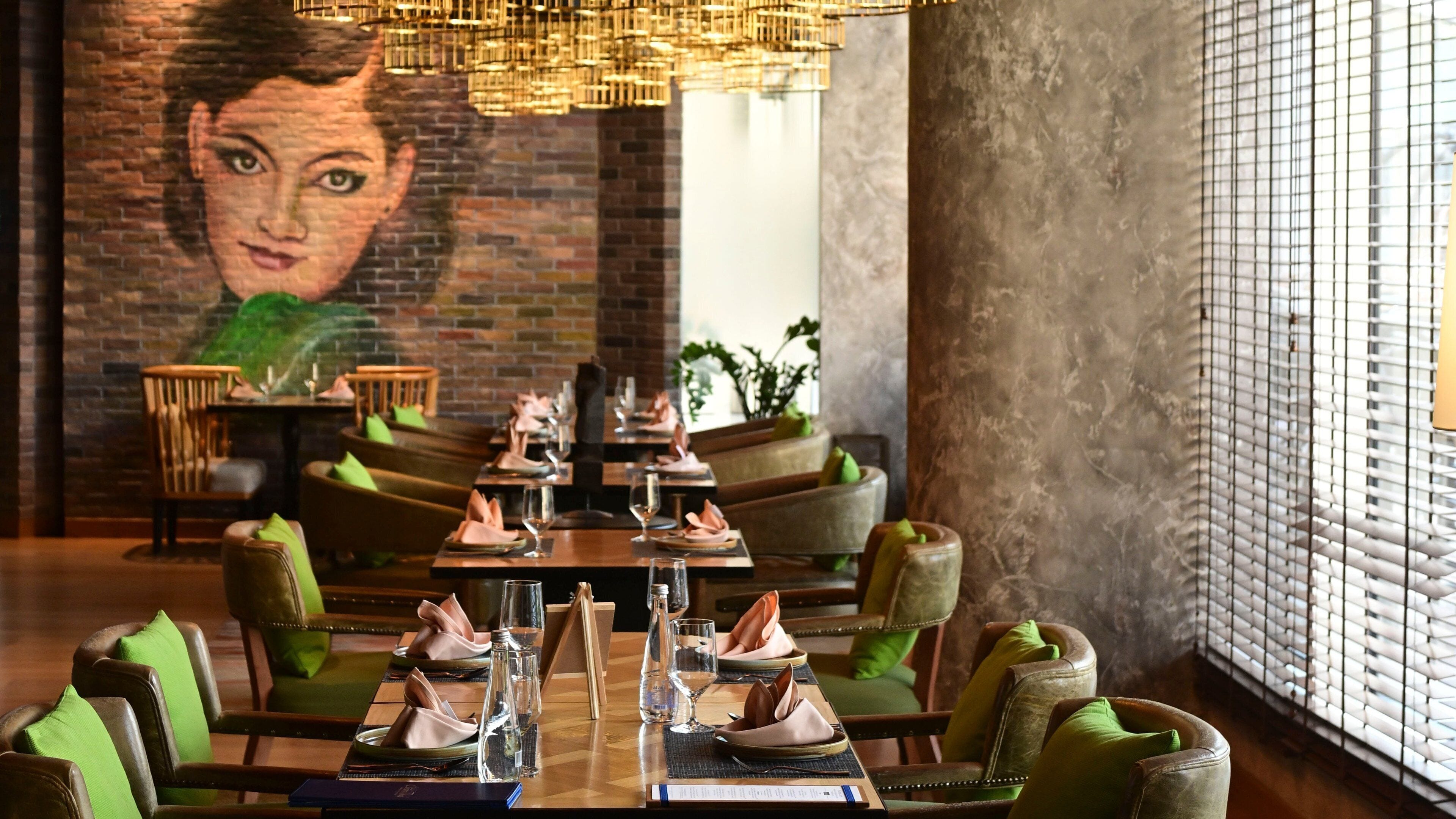 Un restaurant avec une fresque murale en briques, des chaises recouvertes d’un tissu vert et un lustre moderne.