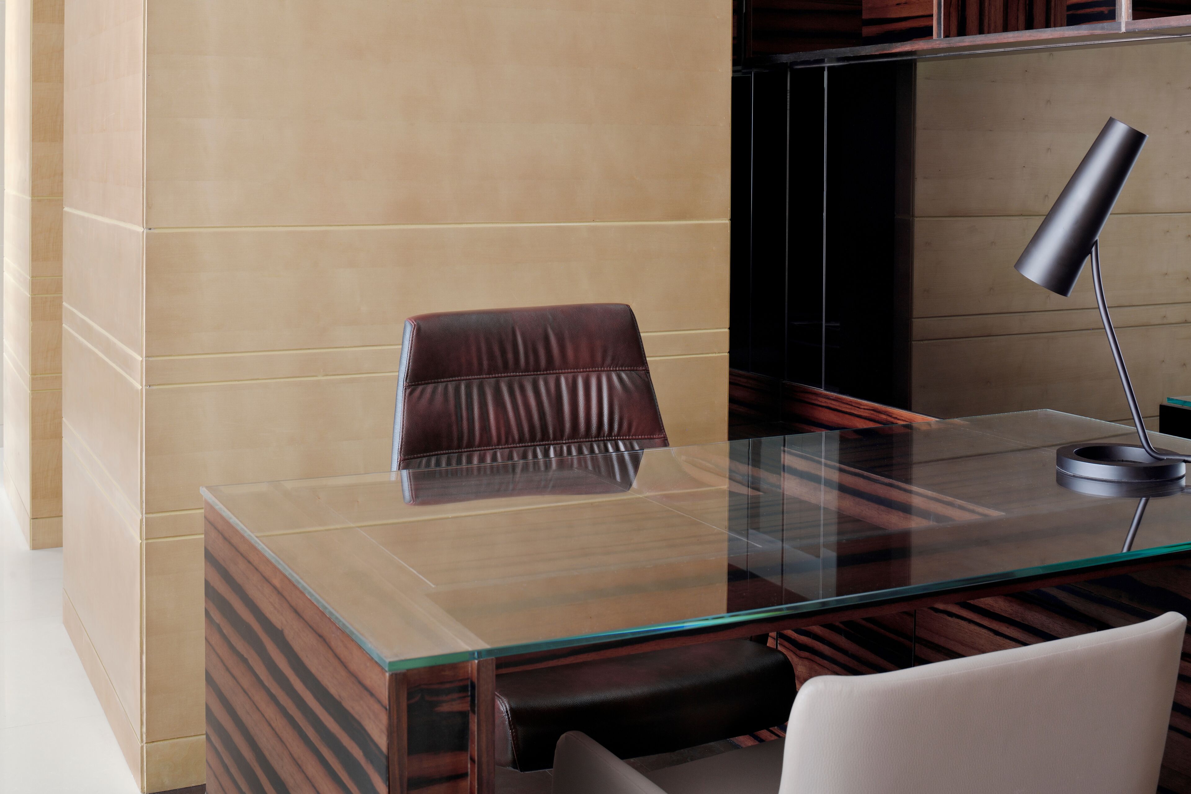 A modern office desk with a leather chair, a glass top, and a black desk lamp.