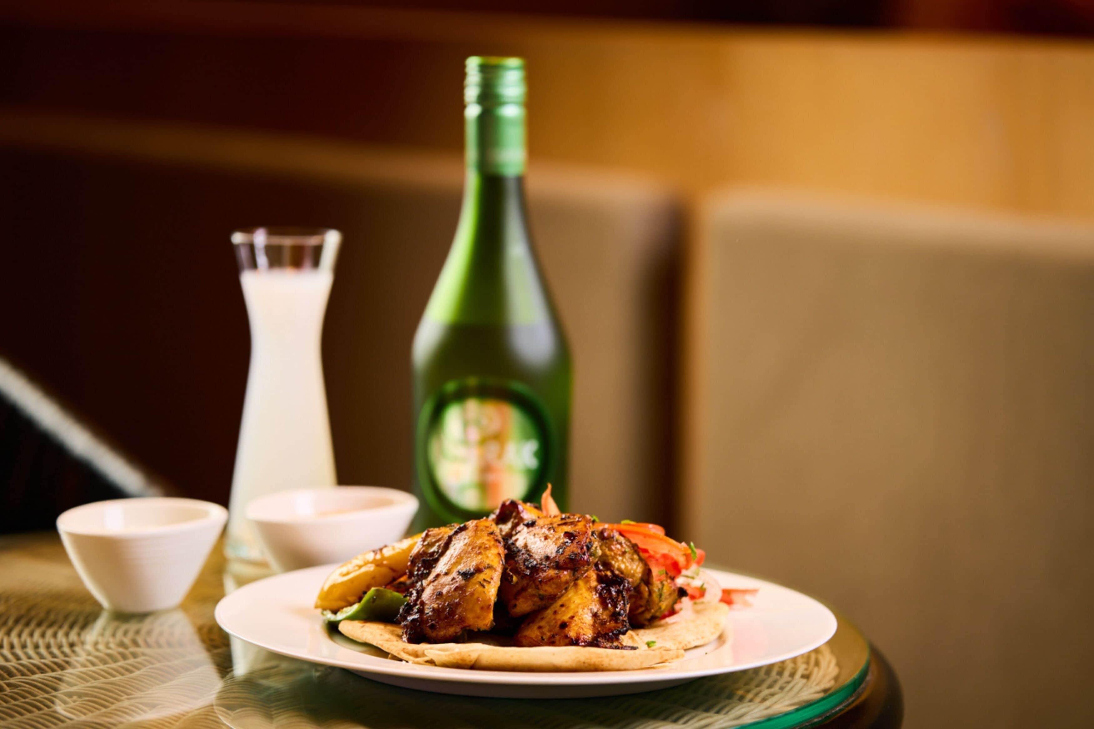 Un plato de pollo a la parrilla con verduras, una botella verde y un vaso de bebida blanca.