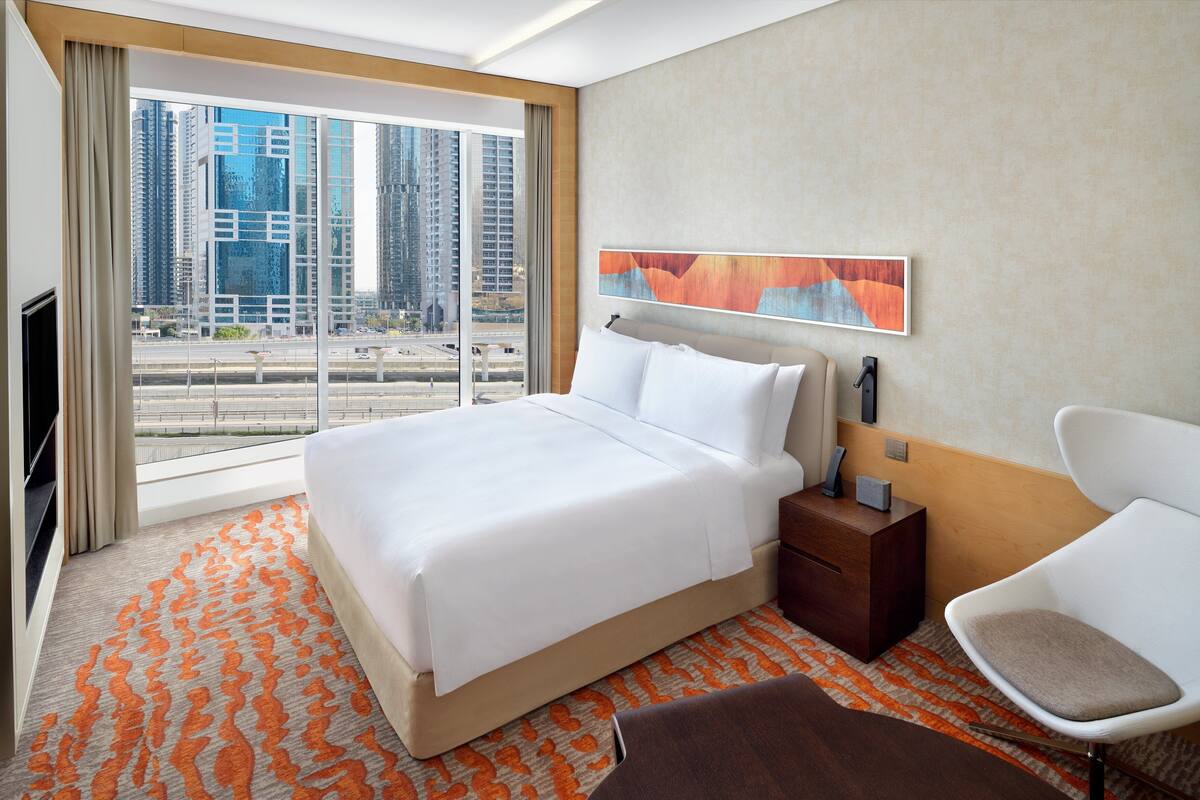 Habitación de hotel con una cama grande, una mesita de noche, una silla y ventana con vista a la ciudad.