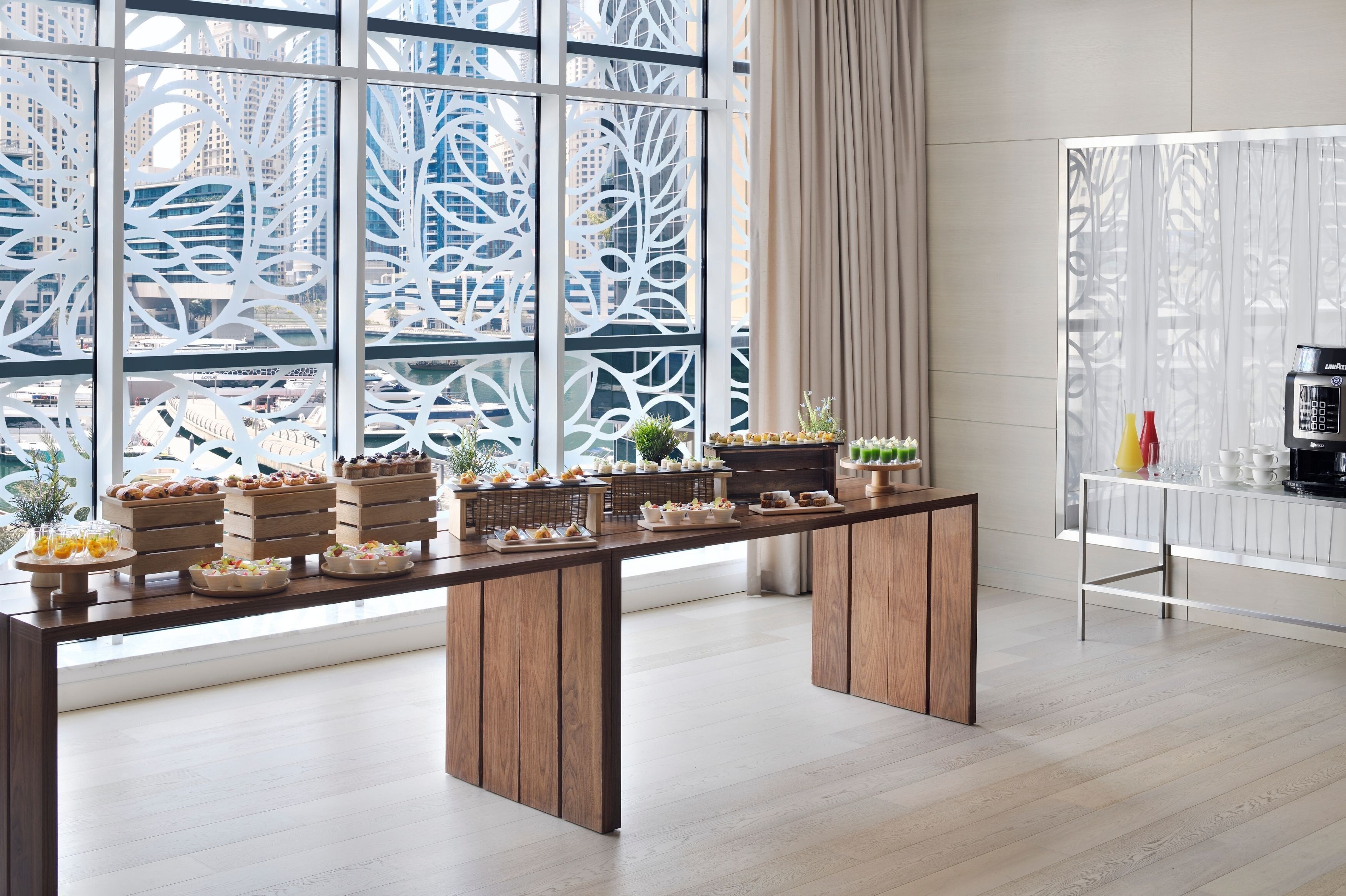 Une table de buffet proposant divers plats et une machine à café, dans une chambre d’hôtel moderne dotée de grandes fenêtres et offrant une vue sur la ville.