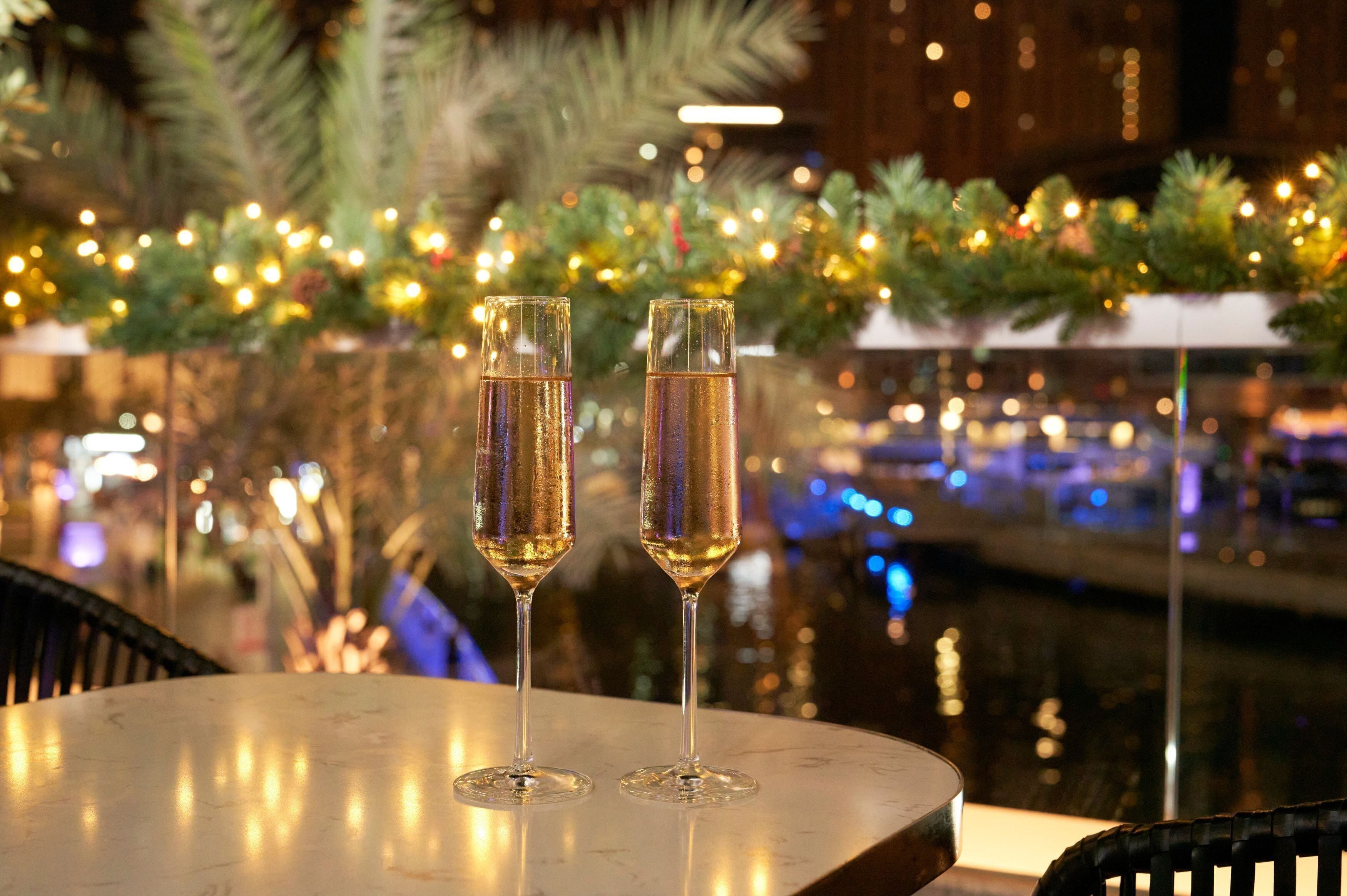 Two champagne flutes on a table with a festive background including greenery and lights.