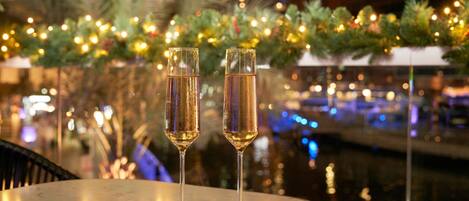 Two champagne flutes on a table with a festive background including greenery and lights.