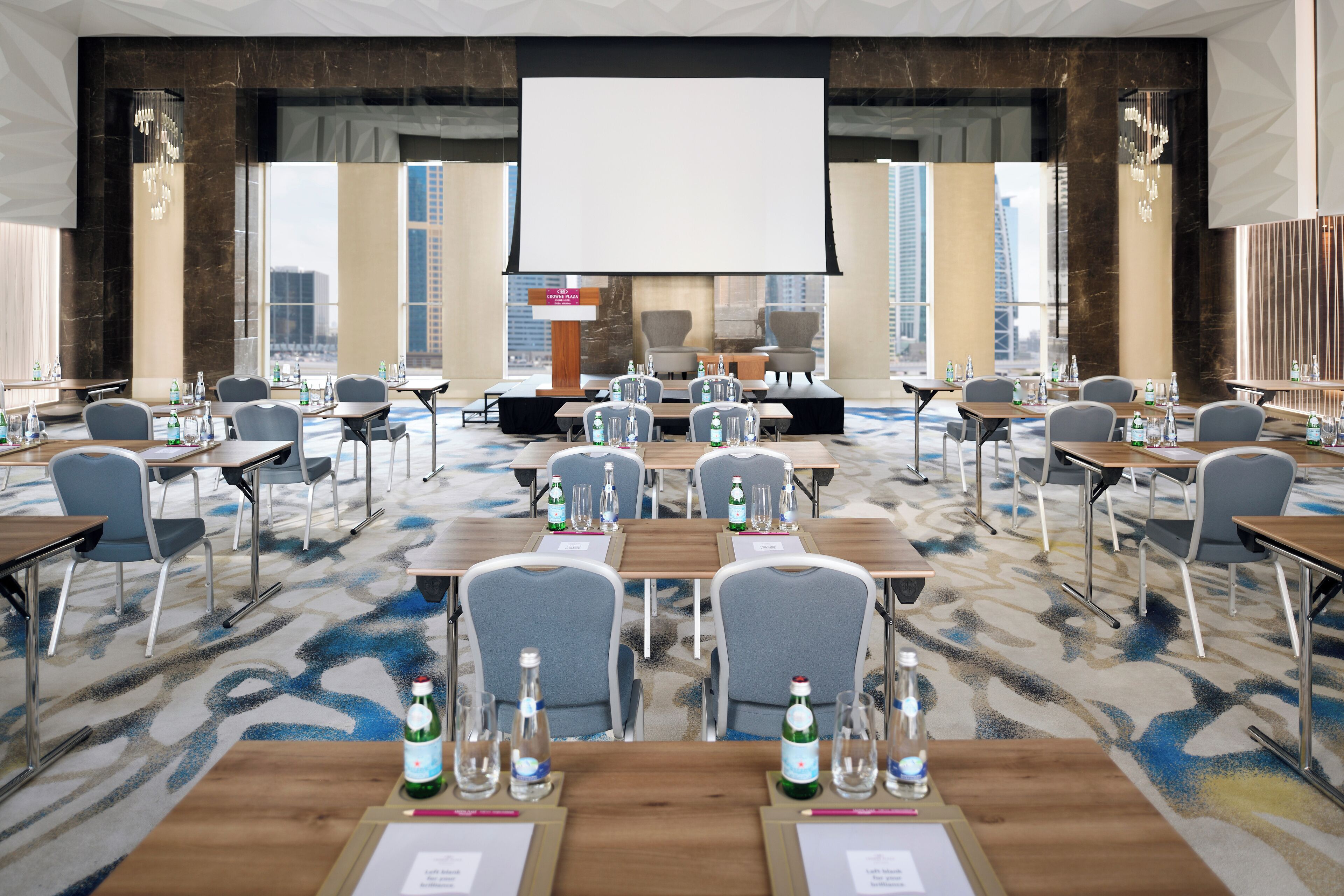 A conference room with a large screen, multiple tables, and chairs, equipped with water bottles and glasses.