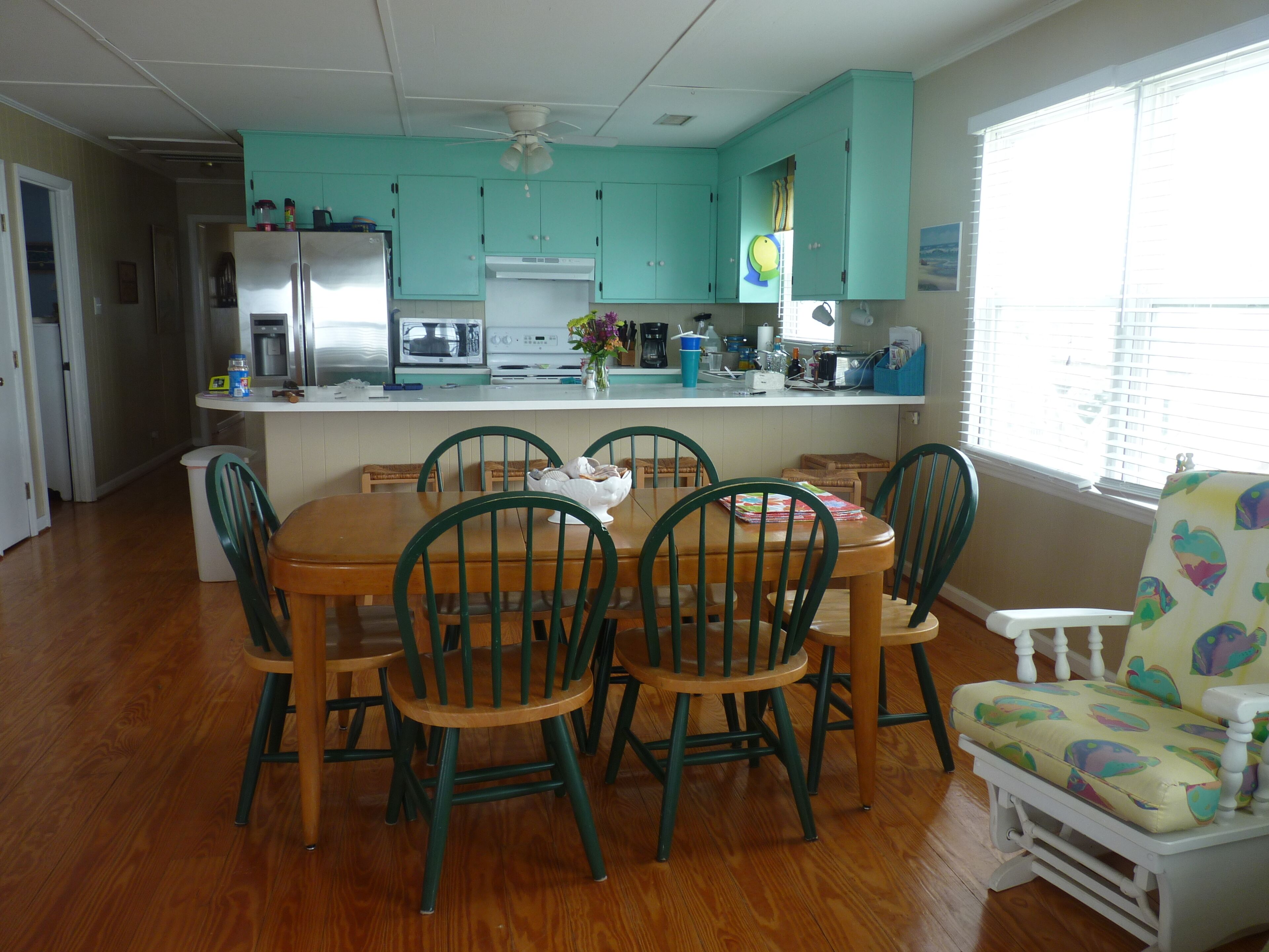 Une cuisine avec des meubles de couleur turquoise, une table à manger avec des chaises et une fenêtre avec des stores.