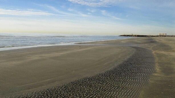 A beach with a wide sandy shore, gentle waves, and a clear sky with scattered clouds.