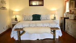 A bedroom with a bed, bedside tables, a lamp, a framed sign, and a wooden bench. - Romantic Ava Haus Cottage. 3 minutes to Main! See our other 4 Ava Haus rentals. (Fredericksburg)