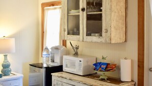 A rustic room with a bed, a white cabinet with glass doors, a microwave, and a small table with a lamp and a bottle of water. - Romantic Ava Haus Cottage. 3 minutes to Main! See our other 4 Ava Haus rentals. (Fredericksburg)