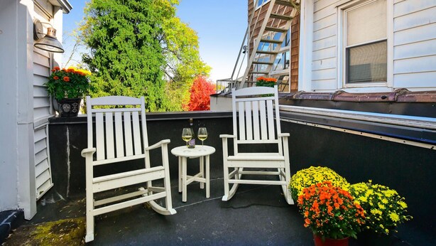 Duas cadeiras Adirondack brancas numa varanda com uma pequena mesa, rodeadas por plantas e flores em vasos.