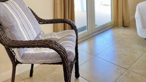 Una silla de mimbre con un cojín a rayas, un espejo redondo en la pared y una puerta corredera de vidrio que da a un espacio exterior.