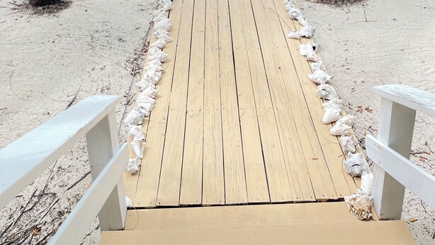 Un sendero de madera que conduce a una zona de piscina, con palmeras y barcos al fondo.