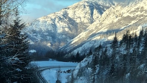 A snow-covered mountain road with a vehicle in the distance, surrounded by evergreen trees and a clear sky. - Girdwood Alaska rental near ski resort. Customers rave: so cute, clean and cozy! (Anchorage)