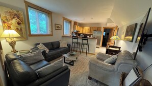 A modern living room with a sofa, a chair, and a coffee table. There is a kitchen area with wooden cabinets and a refrigerator. - Girdwood Alaska rental near ski resort. Customers rave: so cute, clean and cozy! (Anchorage)