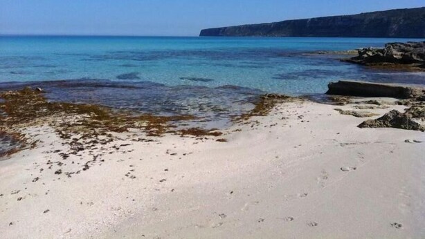 Una playa con aguas cristalinas, afloramientos rocosos y un acantilado lejano.