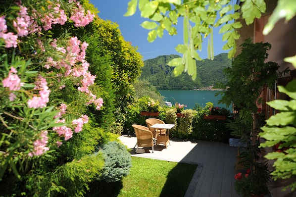 A patio with wicker furniture, surrounded by lush greenery and a view of a lake and mountains.