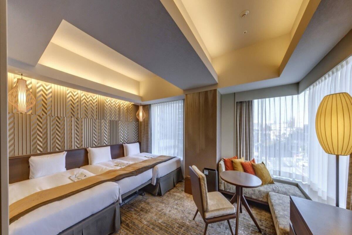 Une chambre d’hôtel avec un grand lit, un canapé, une petite table et une chaise. Il y a une fenêtre avec des rideaux et une vue sur la ville.