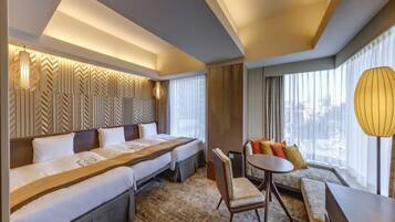 A hotel room with a large bed, a sofa, a small table, and a chair. There is a window with curtains and a view of the city.