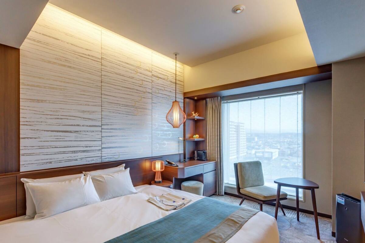 Une chambre d’hôtel avec un grand lit, un bureau, une chaise et une vue sur la ville grâce à de grandes fenêtres.