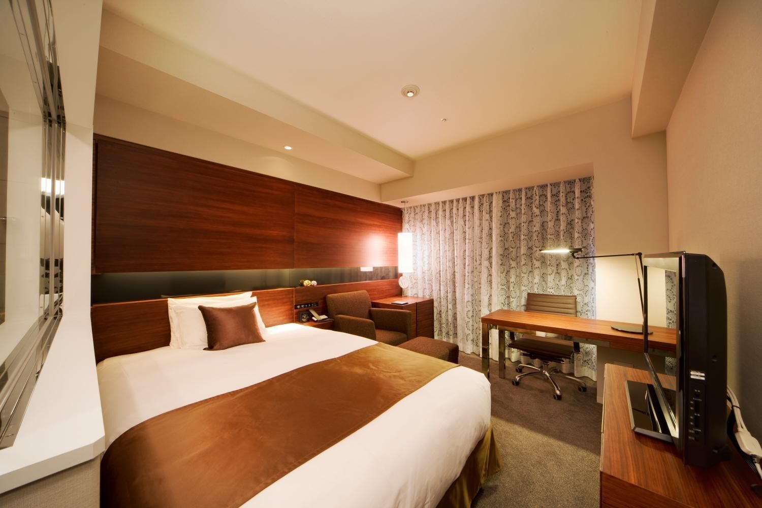 A hotel room with a large bed, a desk, a chair, a TV, and a mirror.