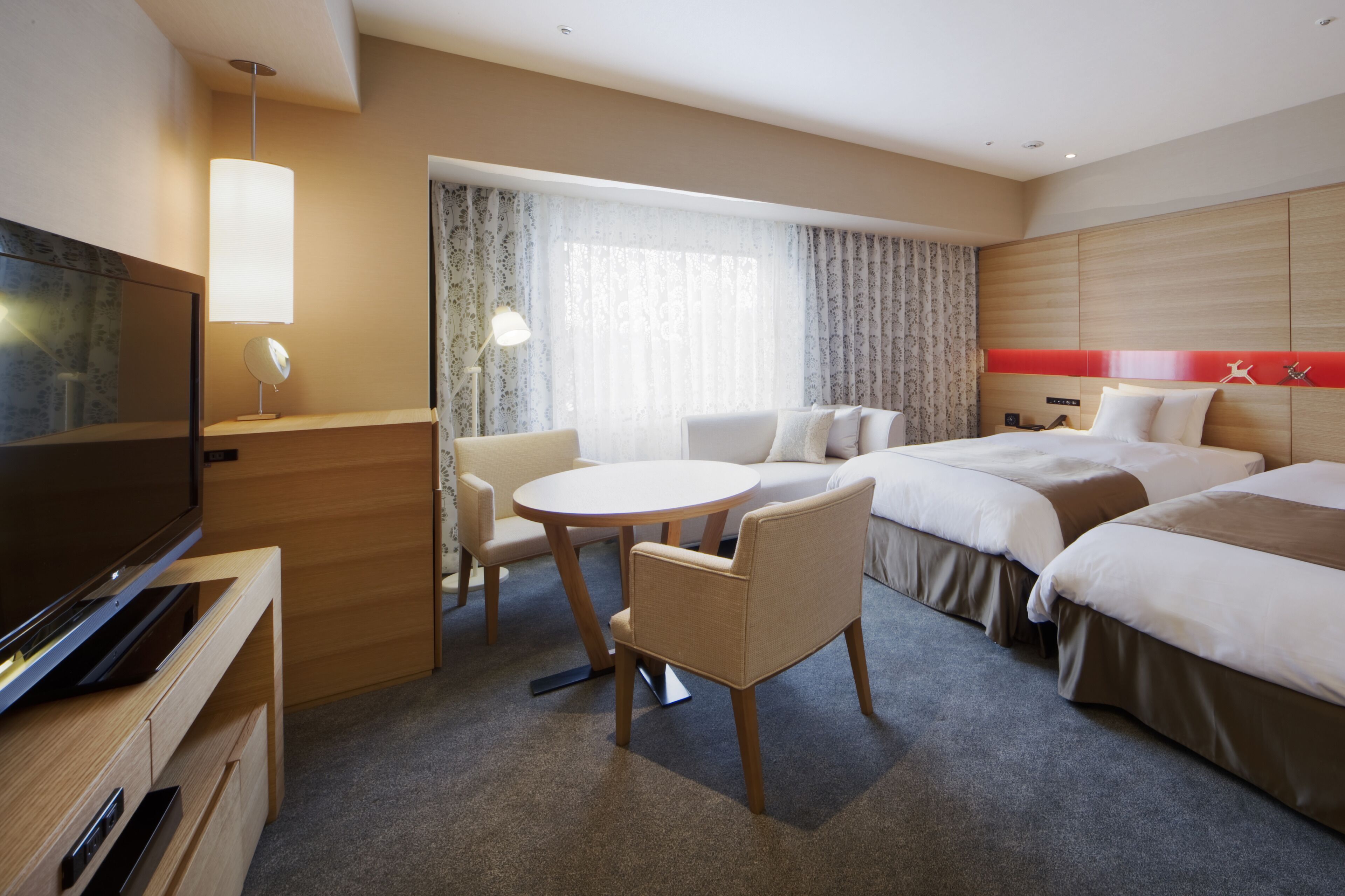 Habitación de hotel con televisión, una mesa redonda, dos camas, una silla y una ventana con cortinas.