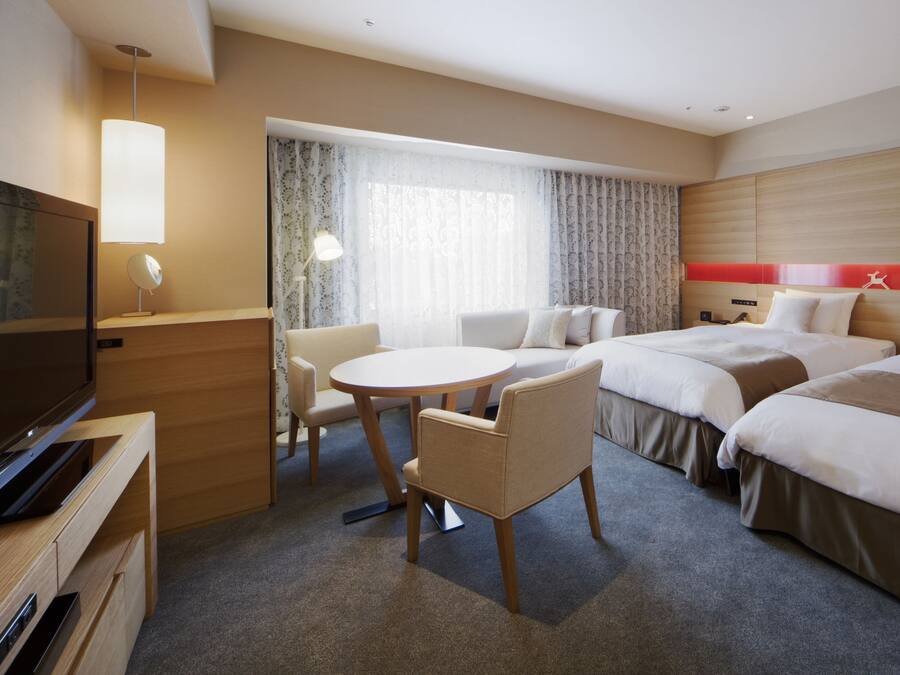 Habitación de hotel con televisión, una mesa redonda, dos camas, una silla y una ventana con cortinas.