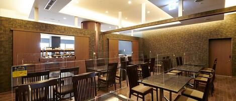 A modern restaurant with glass tables and chairs, a kitchen area in the background, and a high ceiling with recessed lighting.