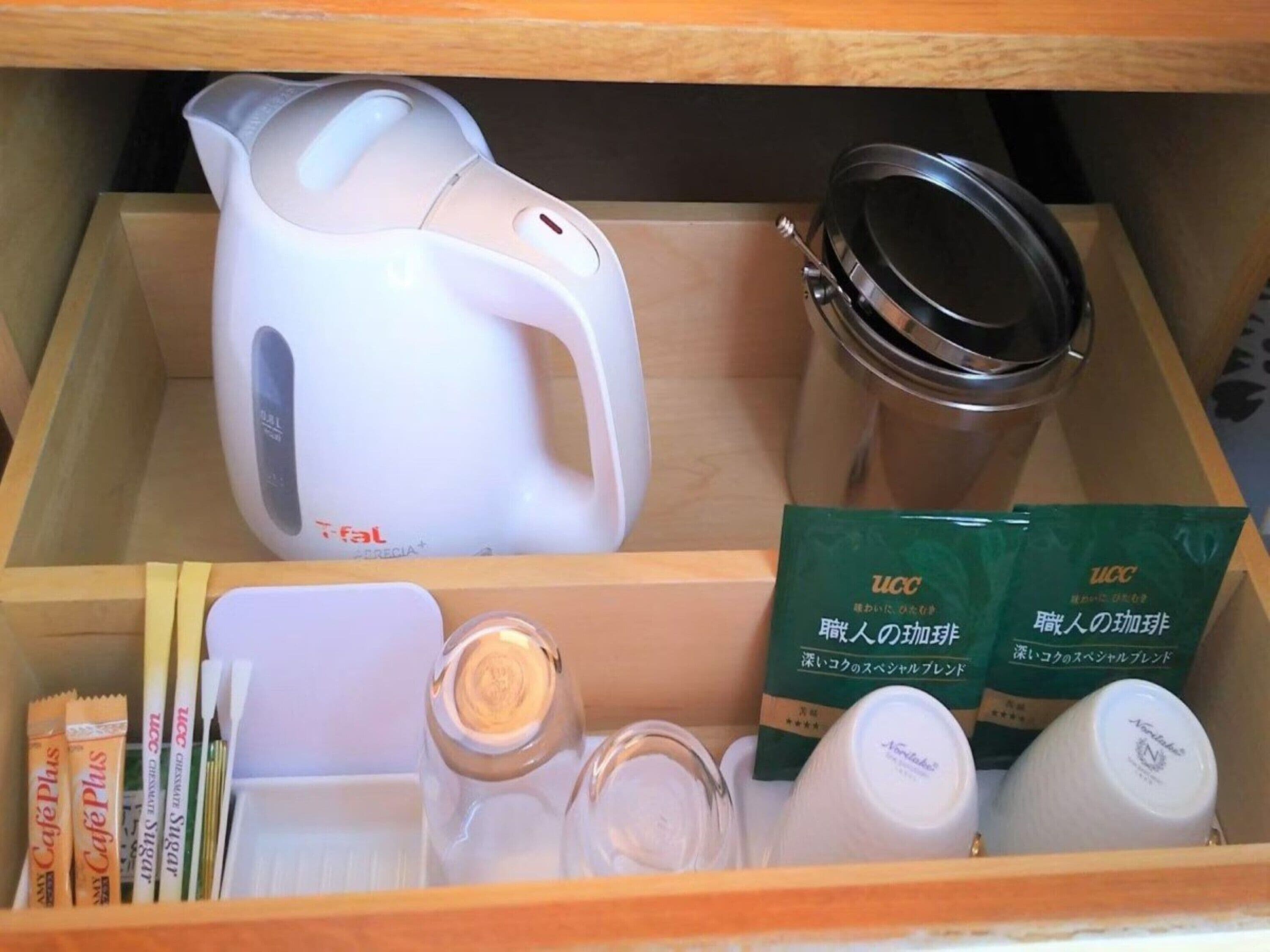 A drawer containing a white electric kettle, a stainless steel container, two green UCC branded boxes, a white bottle, a small glass jar, and some packaged snacks.