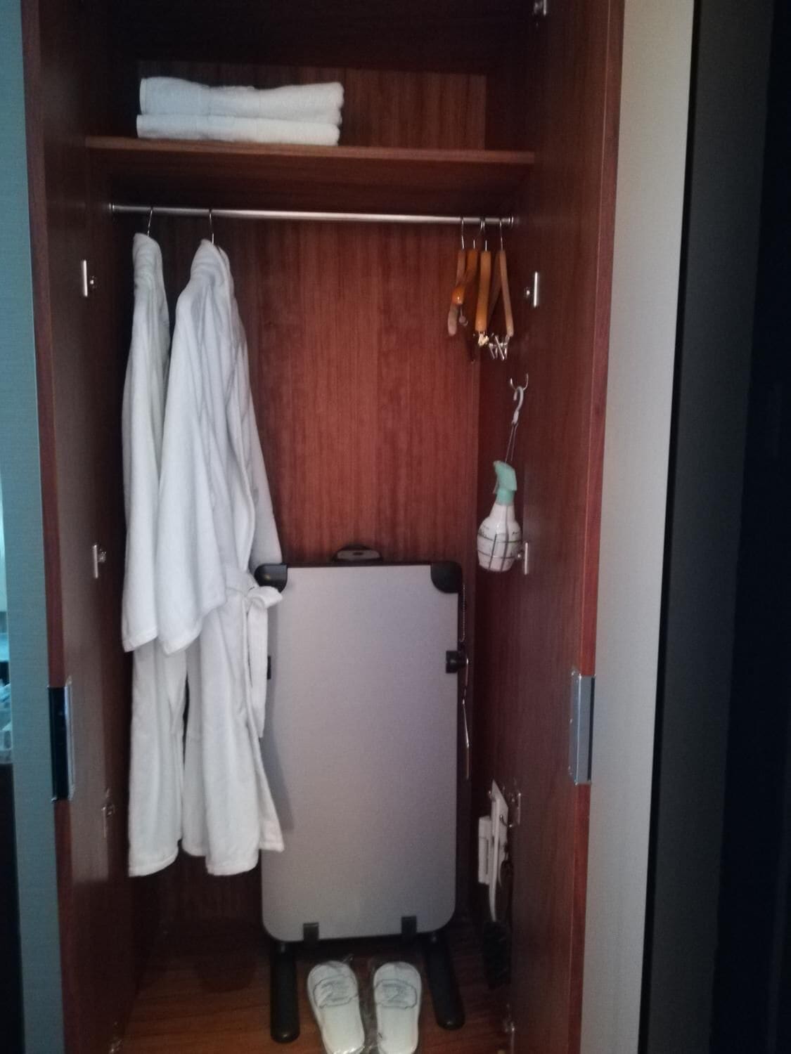 Un armario de madera con dos albornoces blancos colgados en una barra, una toalla blanca en un estante y una pizarra con zapatos en la parte inferior.