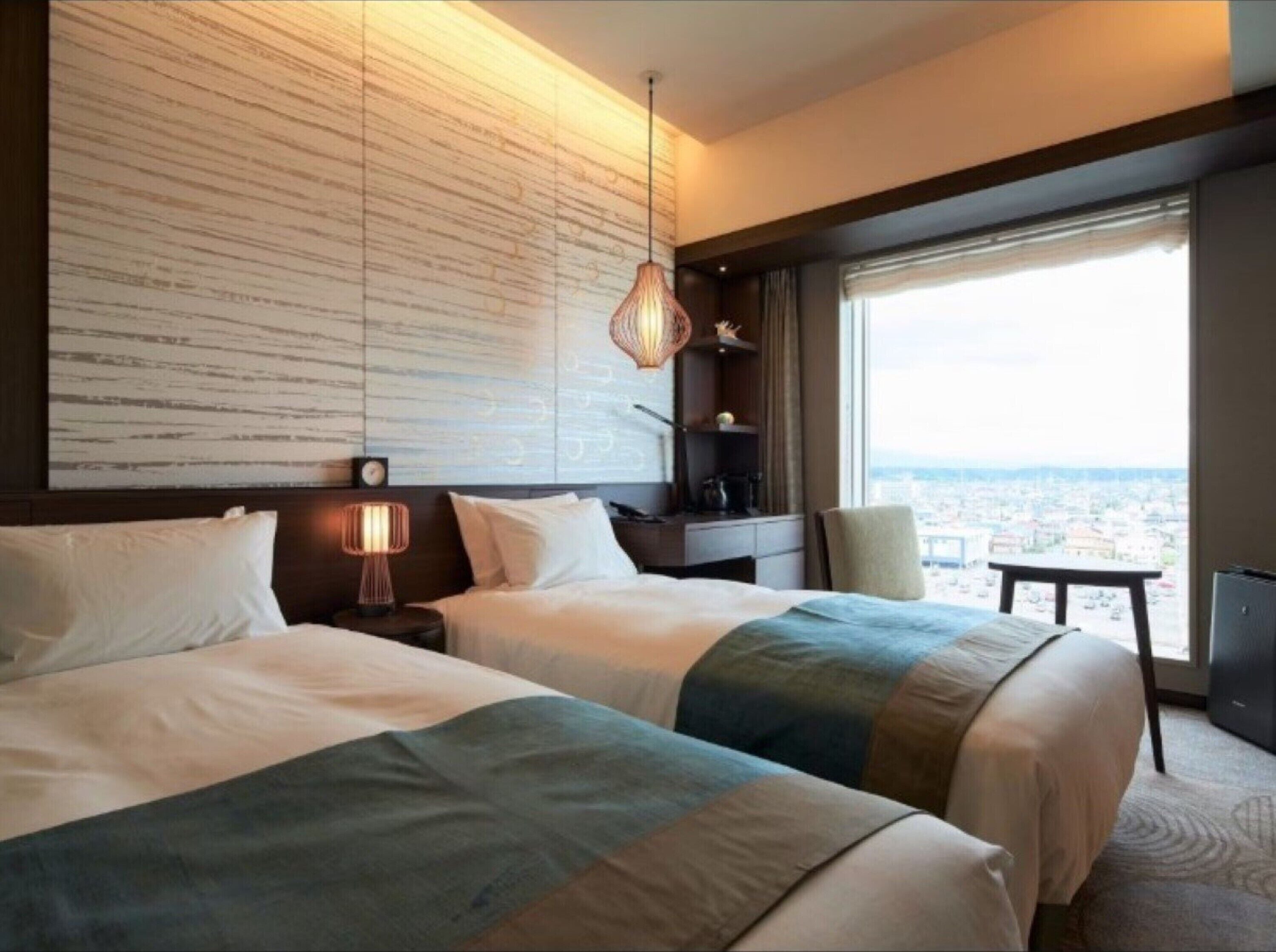A hotel room with two beds, a large window with a city view, a wall with a textured pattern, and a hanging lamp.