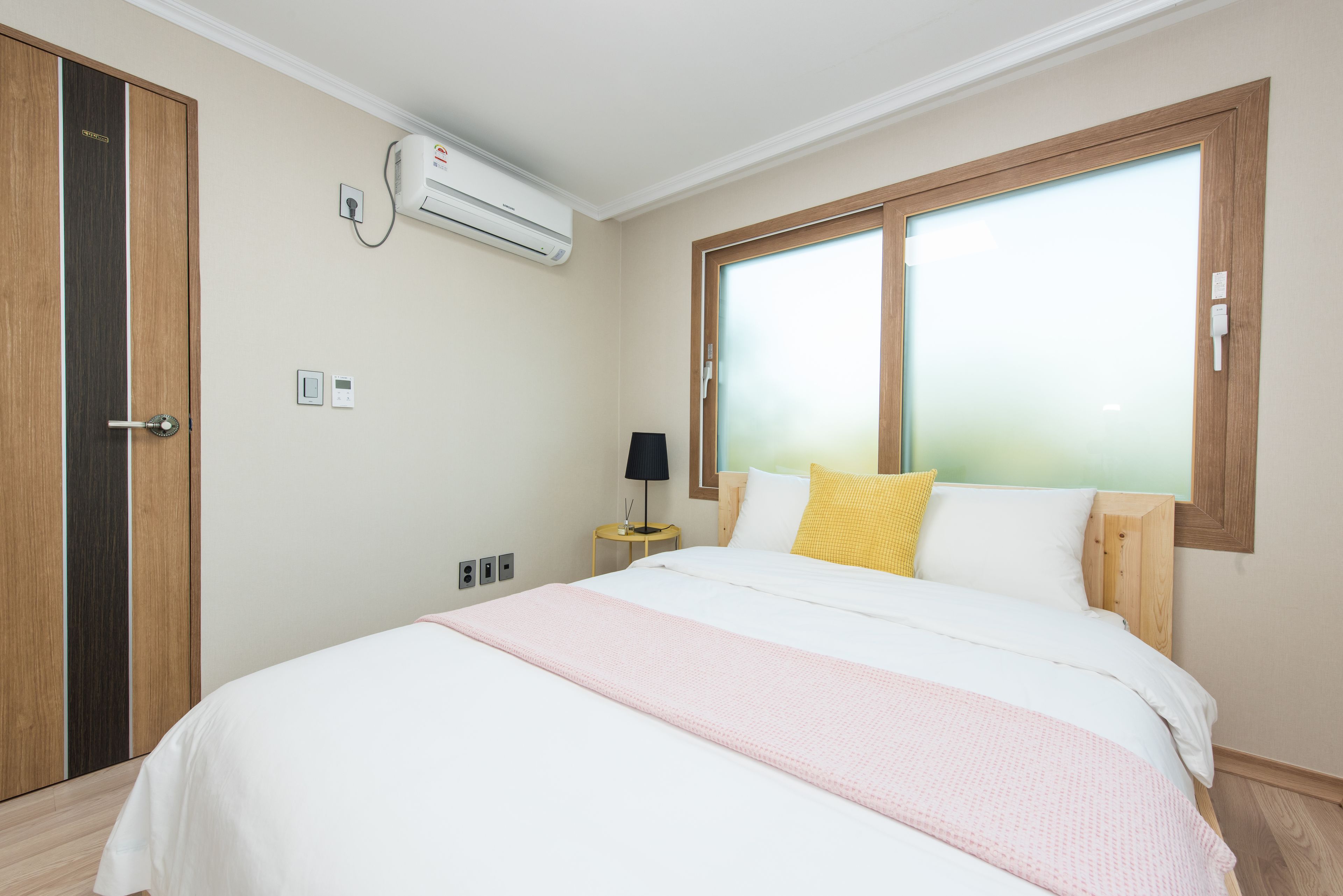 Een slaapkamer met een bed, nachtkastje, lamp, airconditioning en een raam met zonwering.