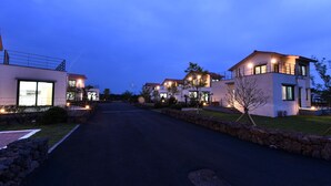 Una calle residencial moderna al anochecer, con casas que tienen amplias ventanas e iluminación exterior.