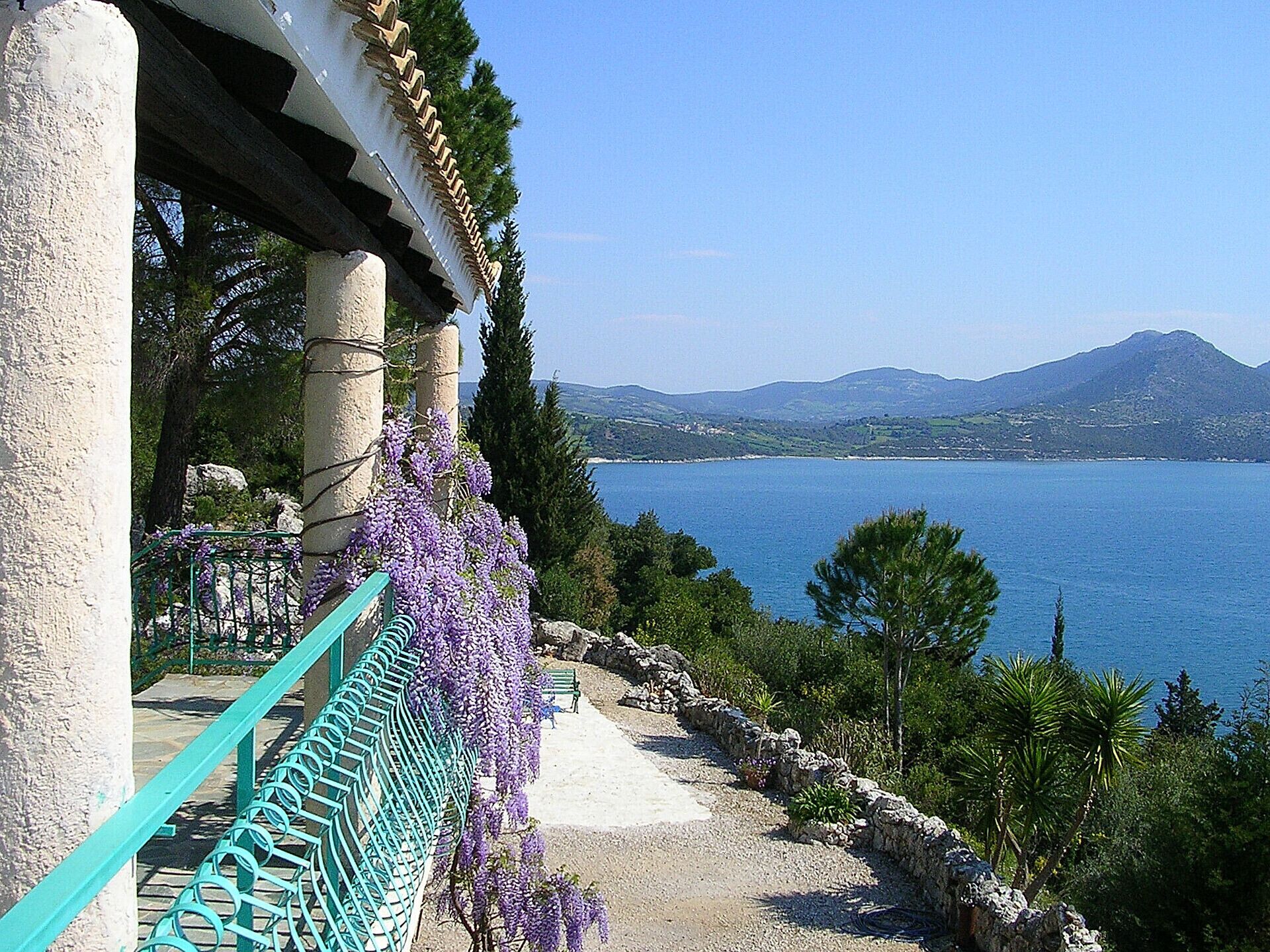 A coastal property with a stone facade, a balcony with purple wisteria, and a view of the sea and mountains.