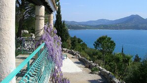 A coastal property with a stone facade, a balcony with purple wisteria, and a view of the sea and mountains.