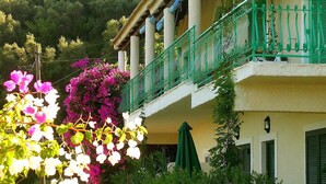 A two-story building with a balcony, surrounded by lush greenery and flowering plants.