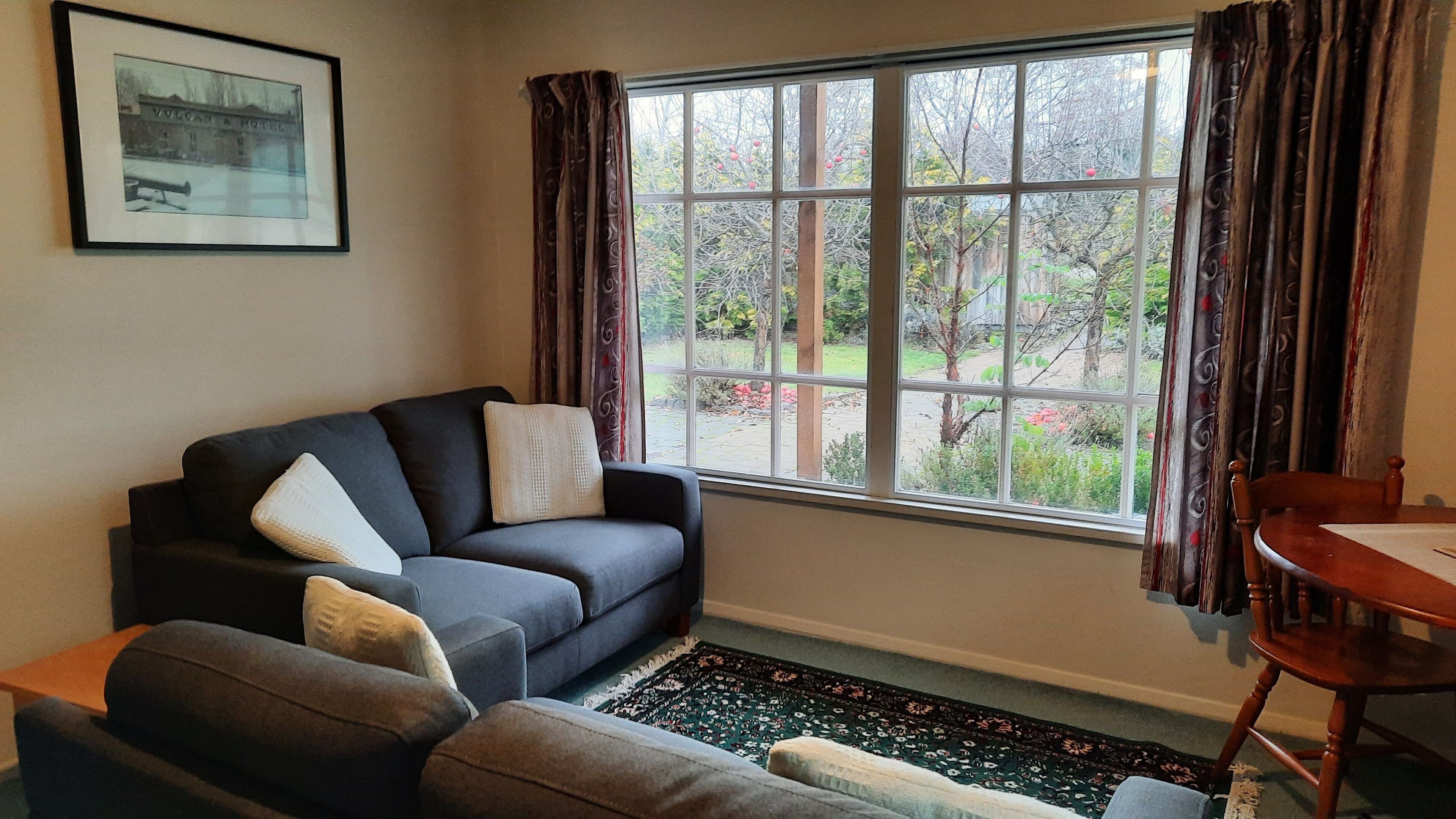 A living room with a dark sofa, a smaller armchair, a wooden table, and a window with curtains.