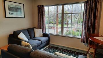 A living room with a dark sofa, a smaller armchair, a wooden table, and a window with curtains.
