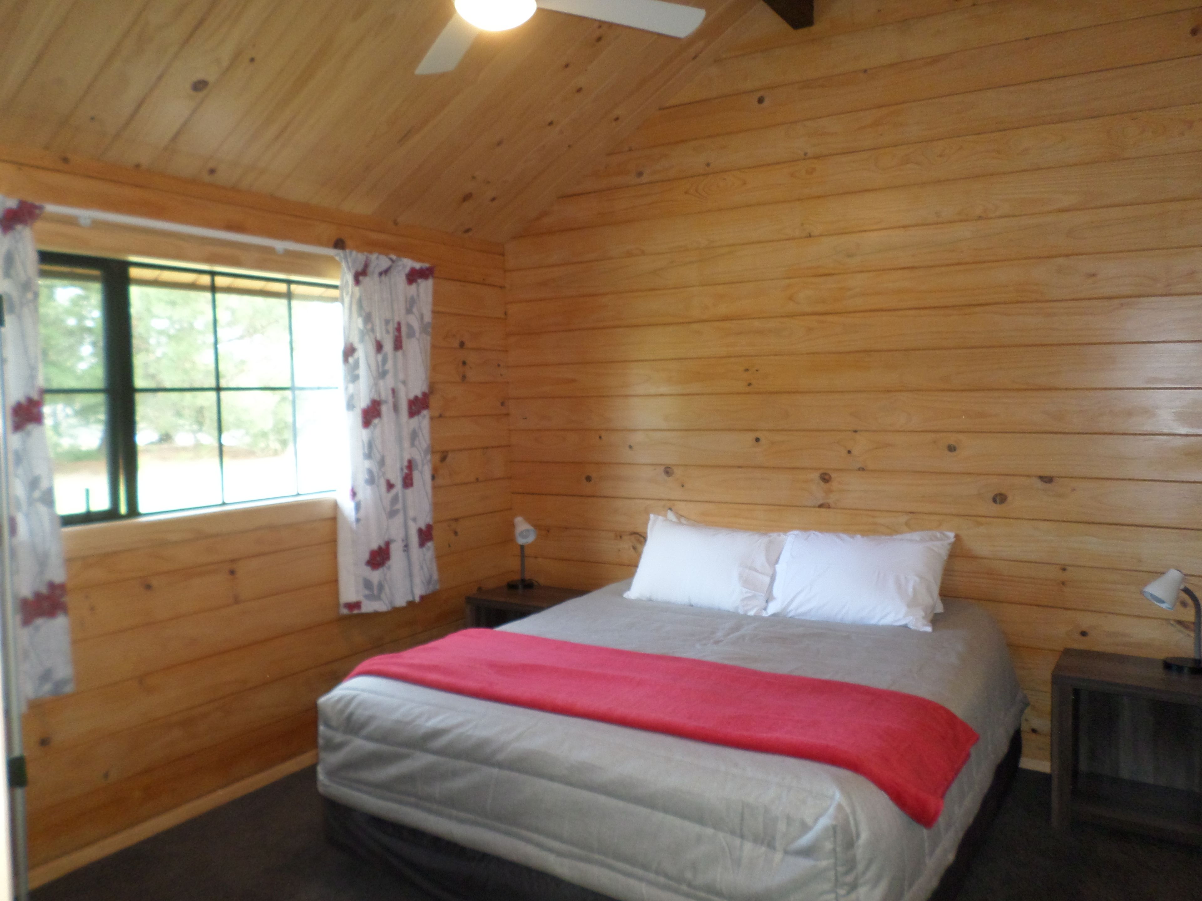 Un dormitorio de cabaña de madera con cama, mesita de noche, lámpara y ventana con cortinas.