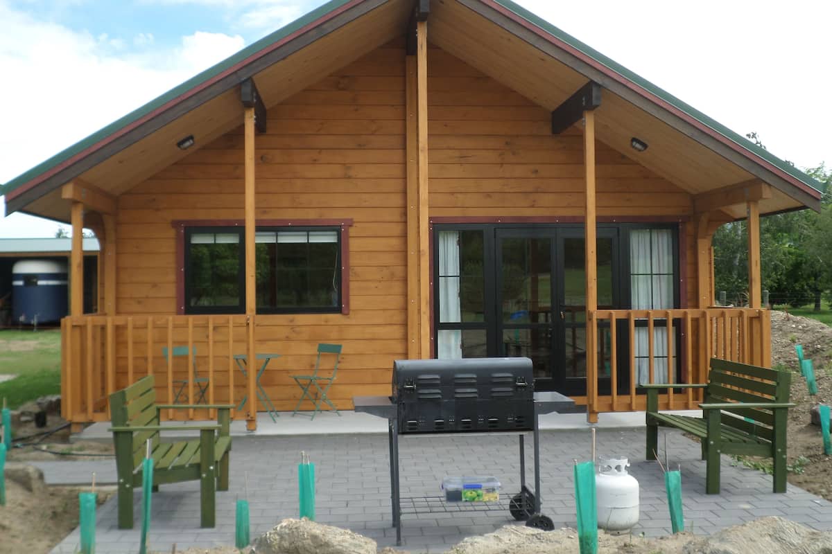 A wooden cabin with a porch, two windows, and a grill outside.