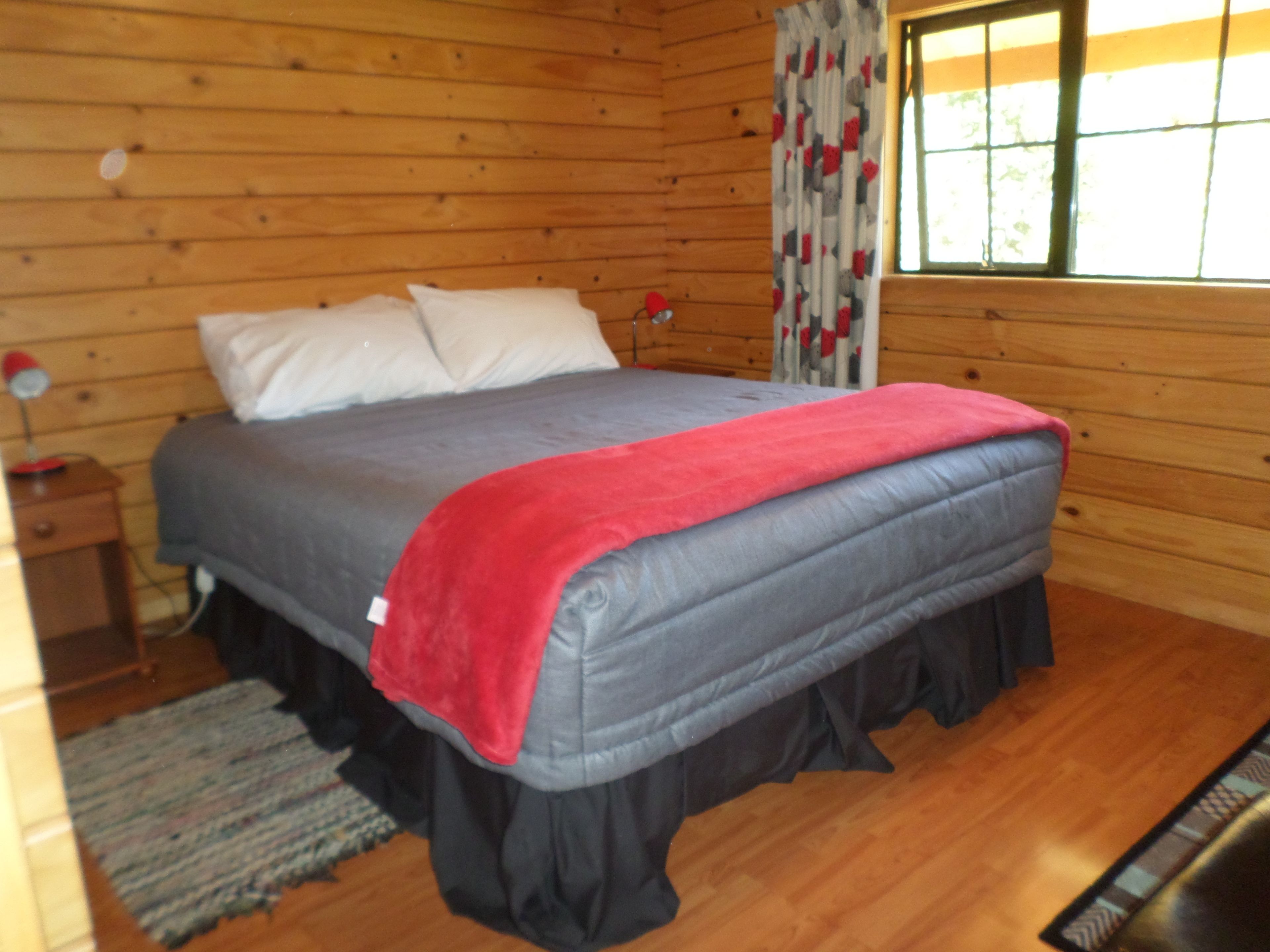 A wooden cabin bedroom with a bed, pillows, a red blanket, a nightstand, a lamp, and a window with curtains.
