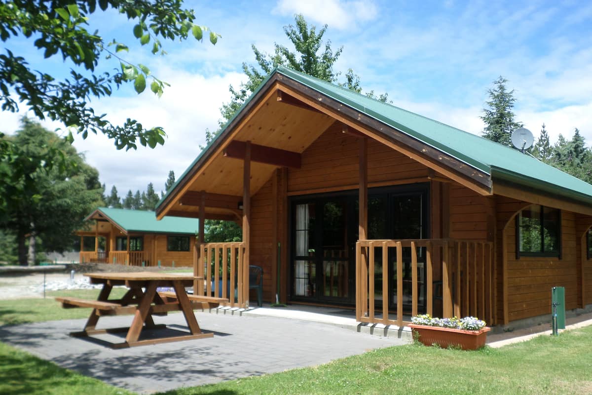 A wooden cabin with a green roof, large windows, and a picnic table in front.