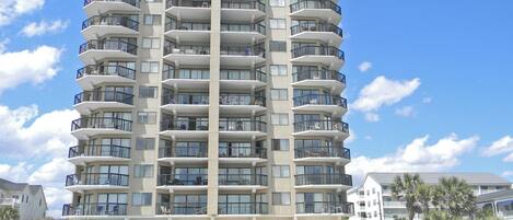 A multi-story hotel with balconies, located near a beach with sand and sparse vegetation.