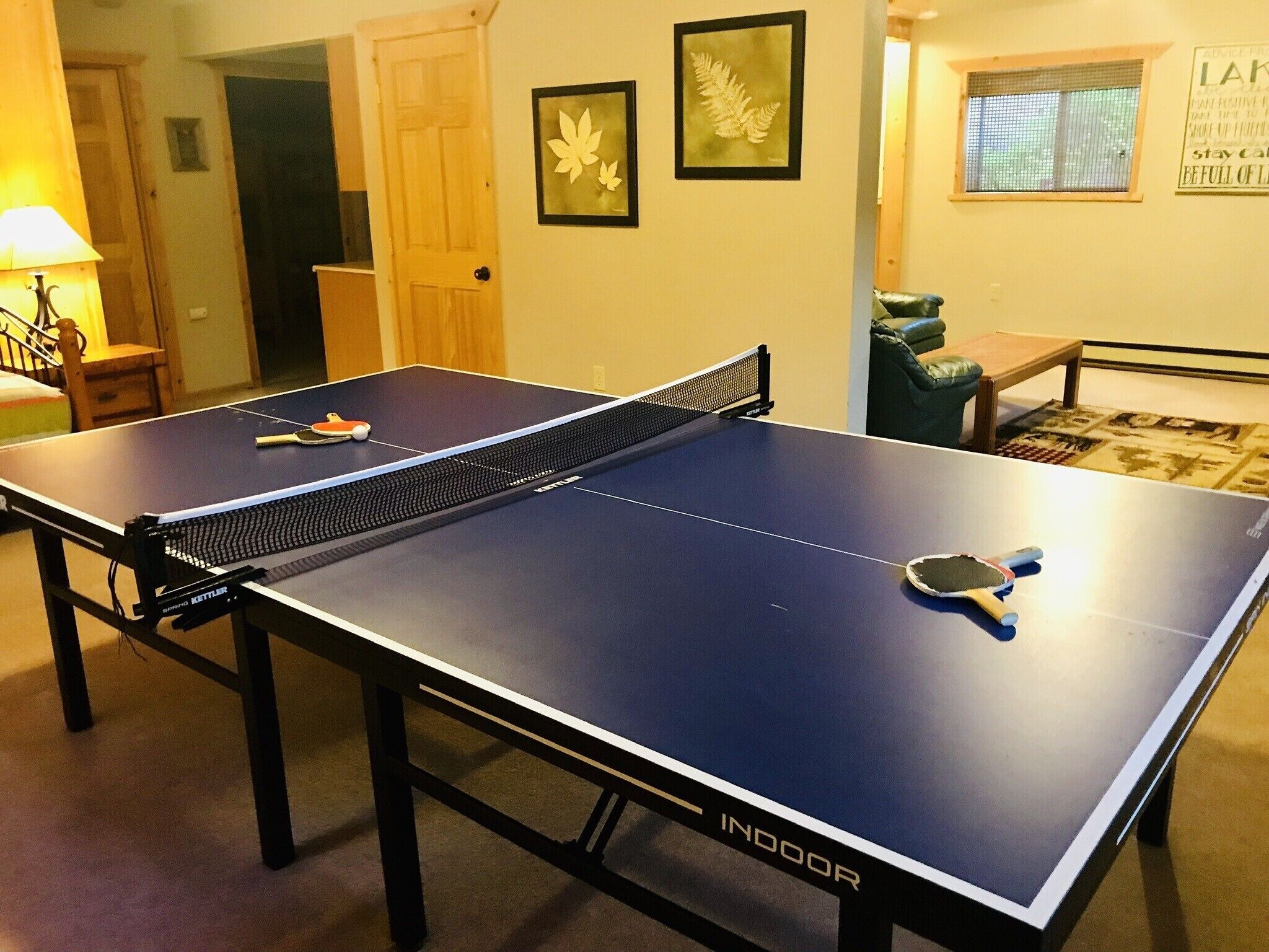 Une table de ping-pong avec un filet, dans une pièce dotée d’une porte, d’une œuvre d’art représentant des feuilles encadrée au mur et d’une fenêtre avec des stores.