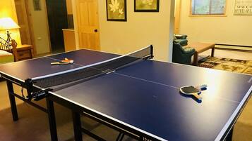 Une table de ping-pong avec un filet, dans une pièce dotée d’une porte, d’une œuvre d’art représentant des feuilles encadrée au mur et d’une fenêtre avec des stores.