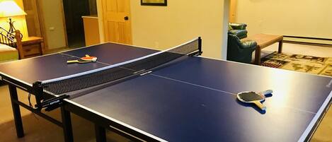 A ping pong table with a net in a room with a door, framed leaf artwork on the wall, and a window with blinds.