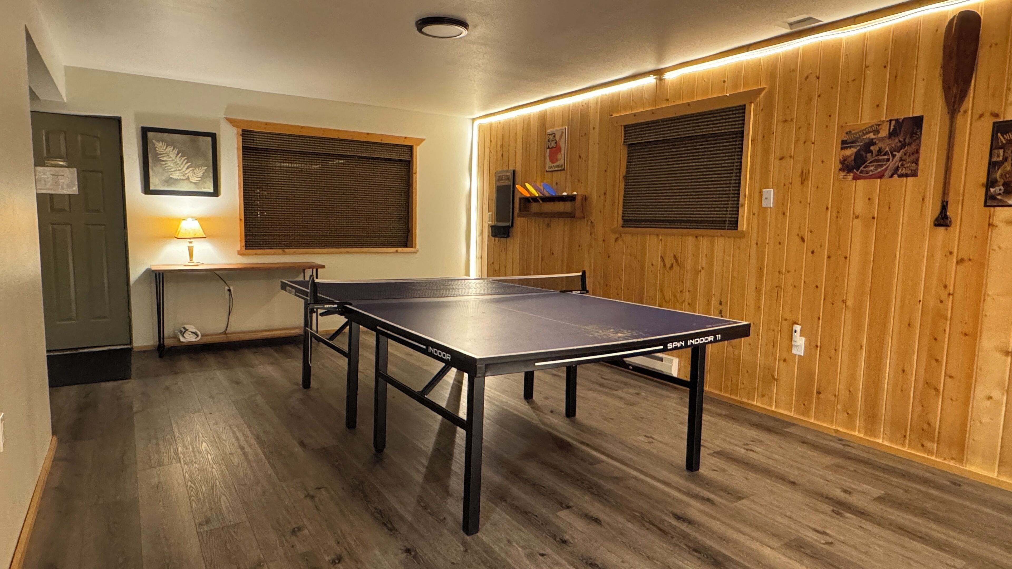 A room with a ping pong table, wooden walls, and a window with blinds.