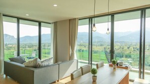 A modern living room with a sofa, dining table, and large windows offering a view of a green landscape. - Sandao Condo Khaoyai (Pak Chong)