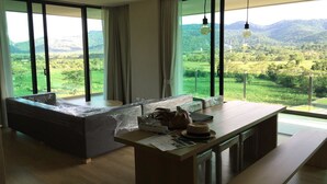A modern living room with a large window, a sofa, a wooden table, and a hanging light fixture. - Sandao Condo Khaoyai (Pak Chong)
