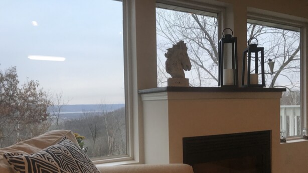 A cozy living room with a fireplace, a sofa with patterned cushions, and a view of trees and a distant landscape through large windows.